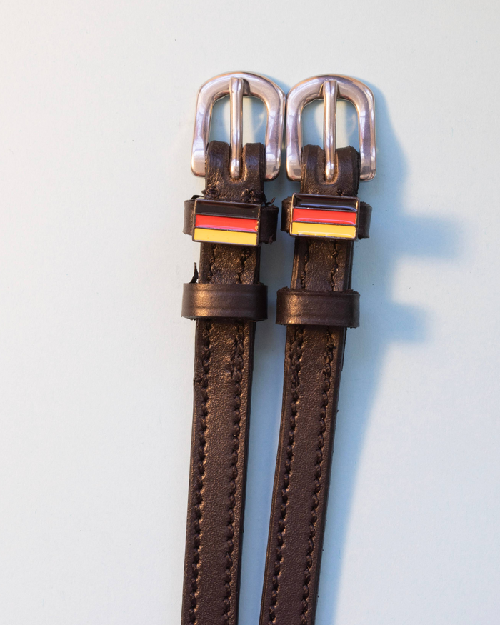 Brown leather ManeJane spur straps with German flag charms and silver buckles on grey