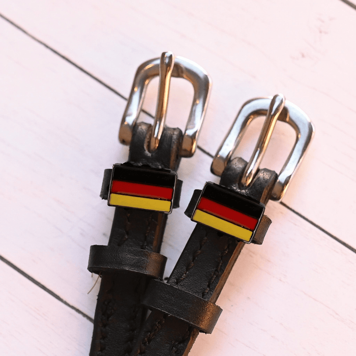 German flag charm spur straps in black leather on a pale pink wood-plank background