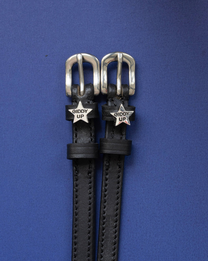 Black leather ManeJane spur straps with silver 'Giddy Up' star charms on a blue background