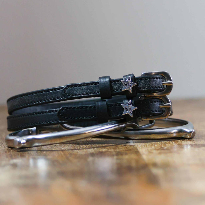 Coiled black leather 'Giddy Up' spur straps stacked on a silver spur on a wooden table