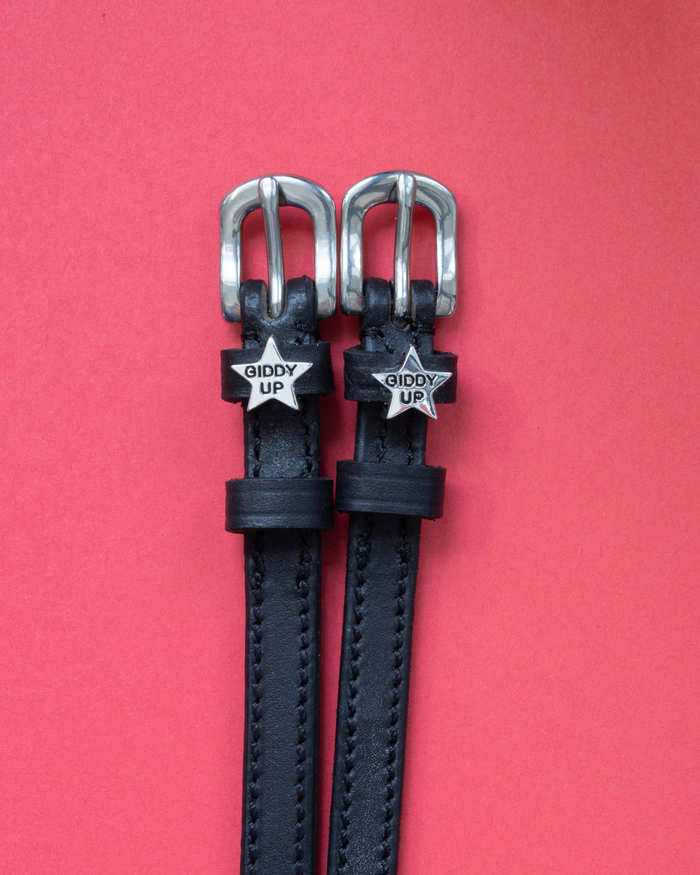 Black leather ManeJane 'Giddy Up' star spur straps with silver buckles on a coral background