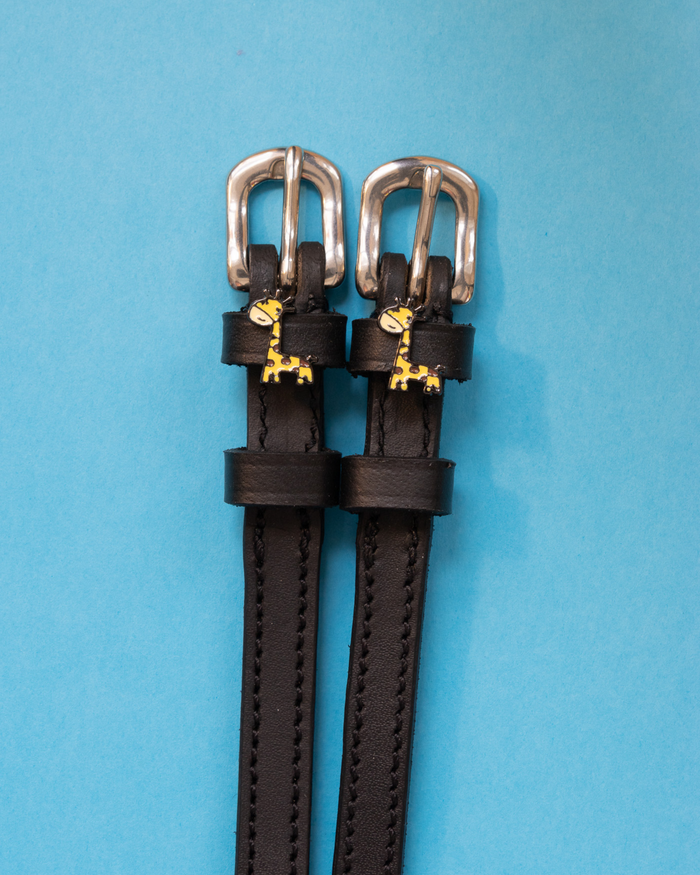 Black leather giraffe charm spur straps with silver buckles on a light blue background