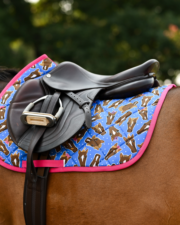 Three-quarter view of the blue Hairy Styles saddle pad beneath a dark leather jump saddle and stirrup
