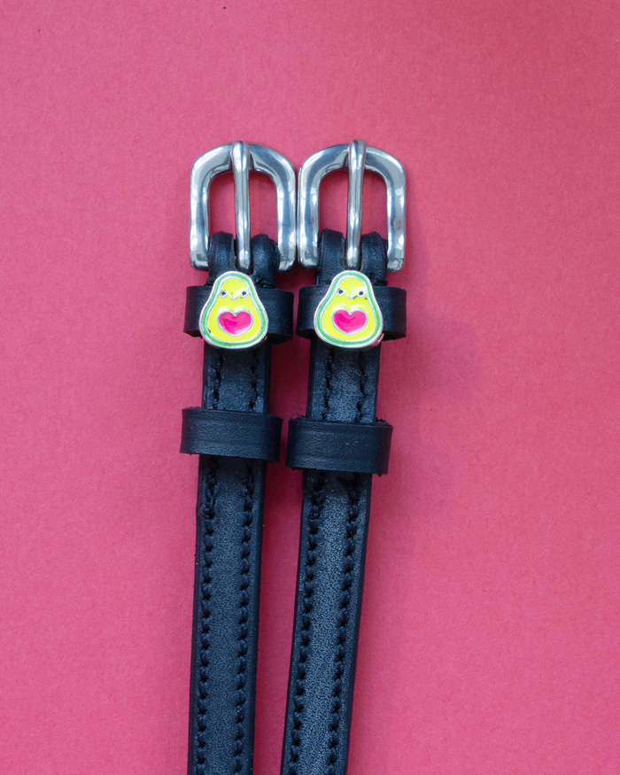 Black leather ManeJane spur straps with heart-pit avocado charms on a pink background