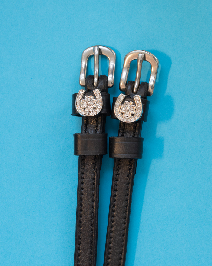 Pair of black leather horseshoe clover spur straps with crystal charms on light blue