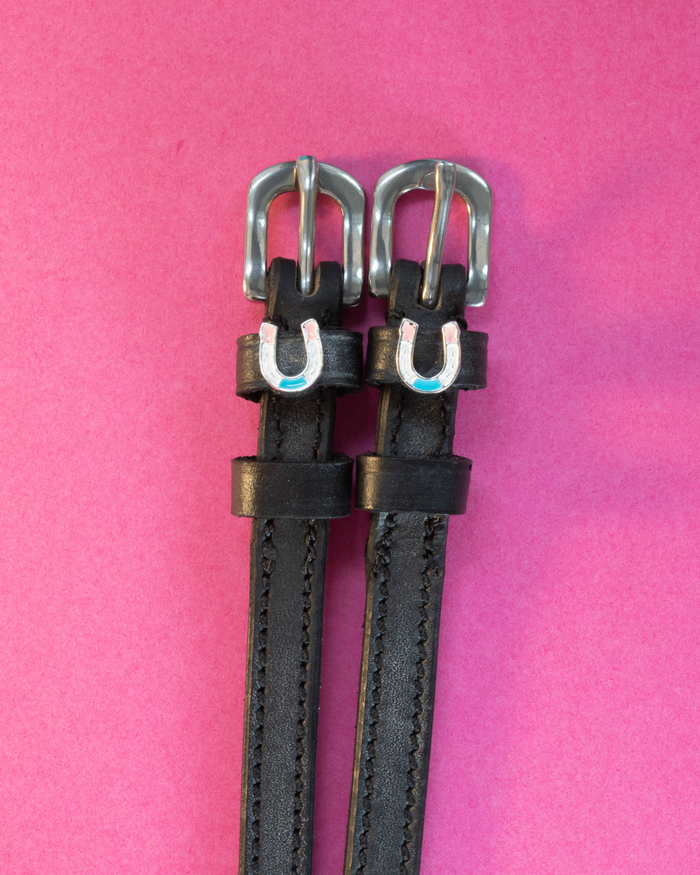 Black leather ManeJane spur straps with pastel horseshoe charms on a hot pink background