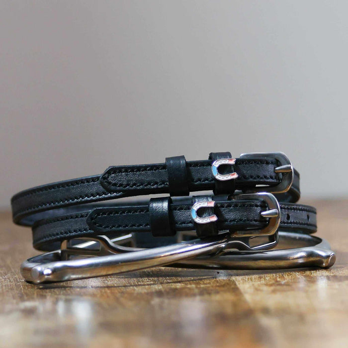 Black leather horseshoe spur straps coiled beside a silver spur on a wood surface