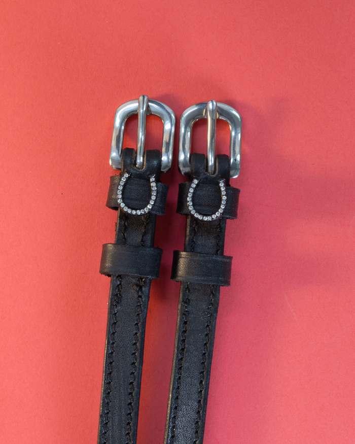 Black leather spur straps with crystal horseshoe charms laid flat on a red background