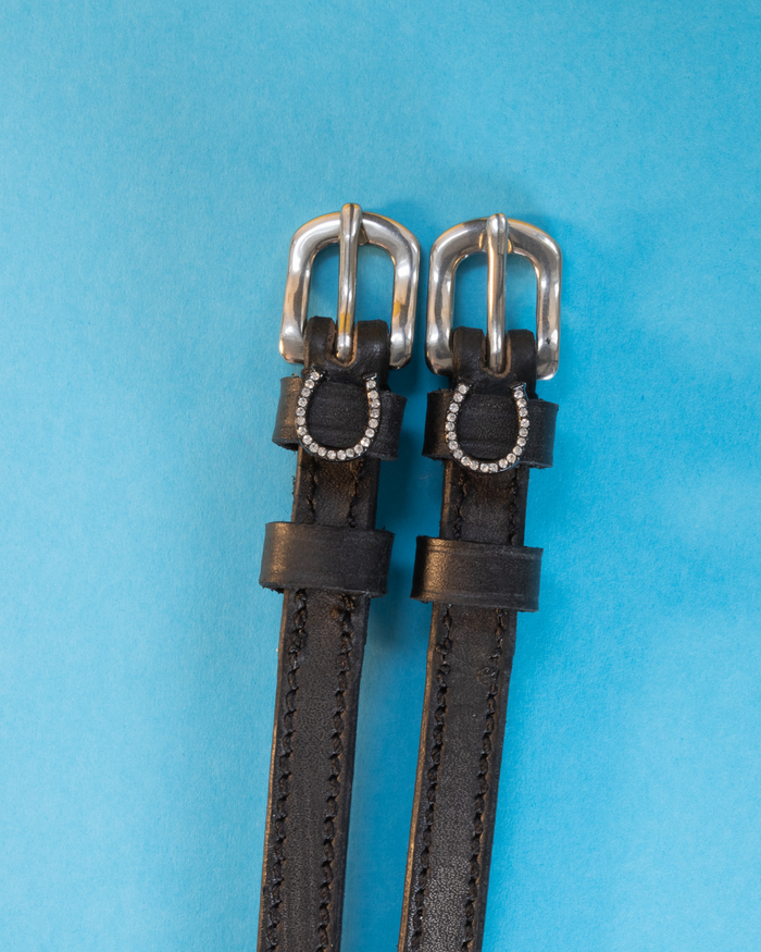 Black leather ManeJane spur straps with rhinestone horseshoes flat on light blue