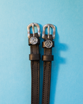 Black leather ManeJane spur straps with silver Leo zodiac charms on a light blue background