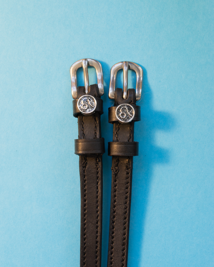Black leather ManeJane spur straps with silver Leo zodiac charms on a light blue background
