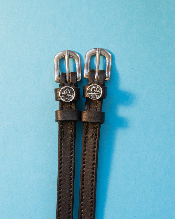 Black leather ManeJane spur straps with round Libra zodiac charms on a bright blue backdrop