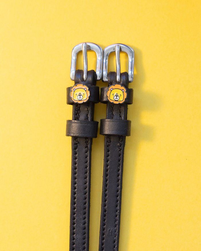 Black leather ManeJane spur straps with cartoon lion face charms and silver buckles on yellow