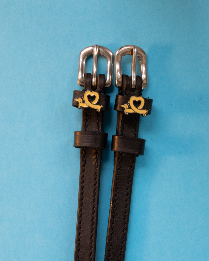 Long Dogs spur straps in black leather with gold dachshund charms on a light blue background