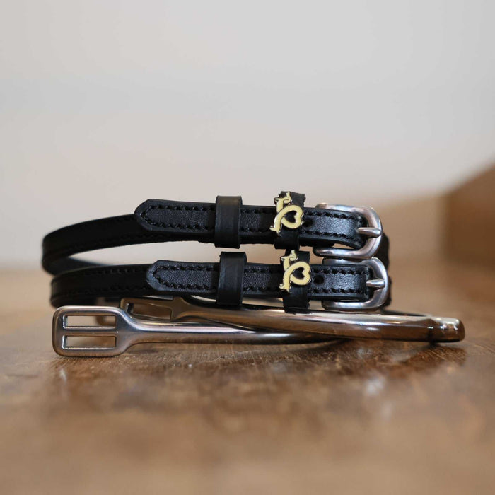 Stacked black Long Dogs spur straps with silver buckles on wood beside a stirrup iron, ManeJane