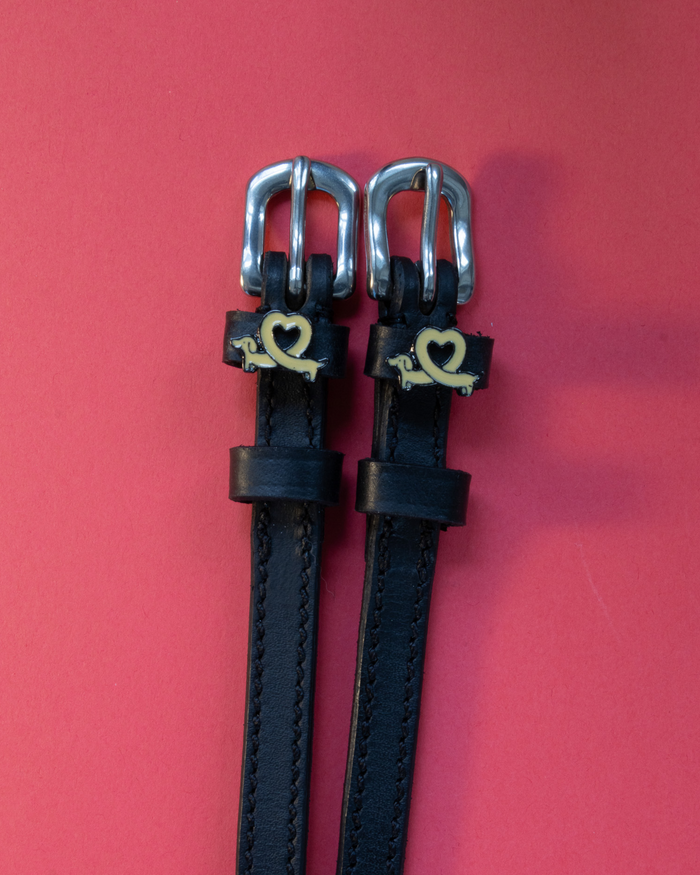 Black leather Long Dogs spur straps with gold dachshund charms on a coral background