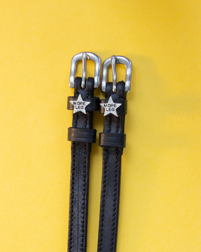 Pair of black leather MORE LEG star spur straps flat-laid on yellow