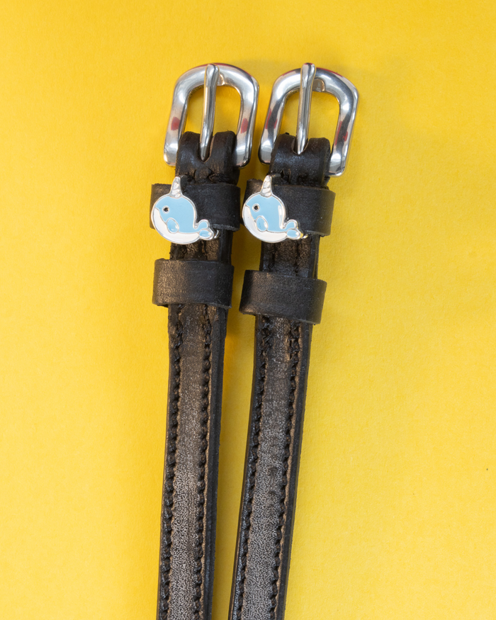 Black leather ManeJane spur straps with blue enamel narwhal charms on a yellow background