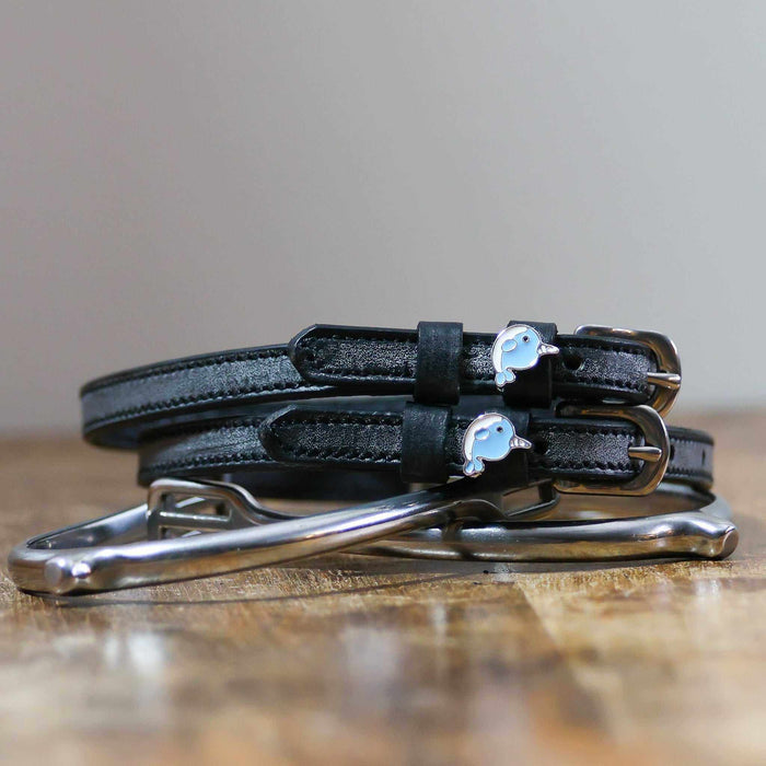 Black leather narwhal spur straps coiled on a wooden surface beside a silver bit