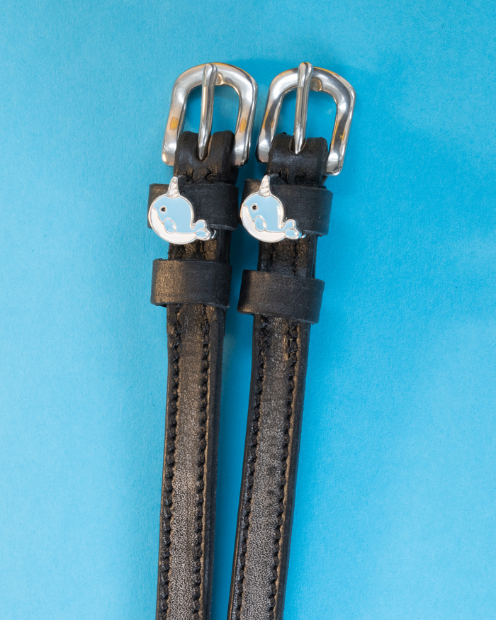 Blue narwhal charm spur straps in black leather laid flat on a light blue background