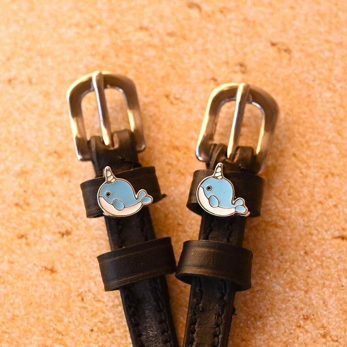 Black leather narwhal spur straps with brass buckles on a sandy cork background