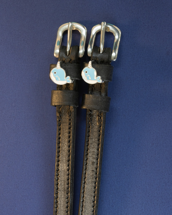 Black leather narwhal charm spur straps flat-laid on a navy blue background