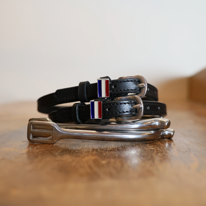 Black leather Netherlands flag spur straps coiled beside a stirrup on a wooden surface