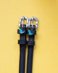 Black leather ManeJane spur straps with teal dinosaur charms on a yellow background