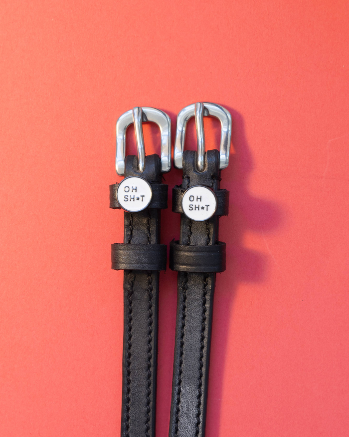 Pair of OH SH*T charm spur straps flat-laid on a coral-red background
