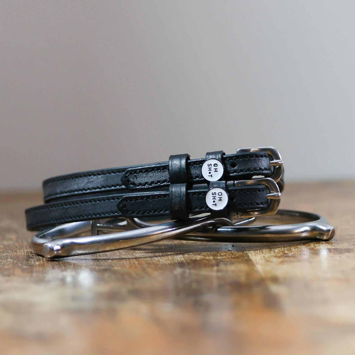 Black leather OH SH*T spur straps coiled beside a stirrup on wood