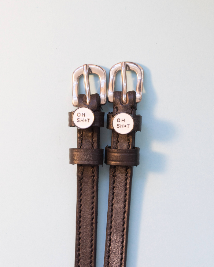 Brown leather ManeJane spur straps with round OH SH*T charms, pale blue backdrop