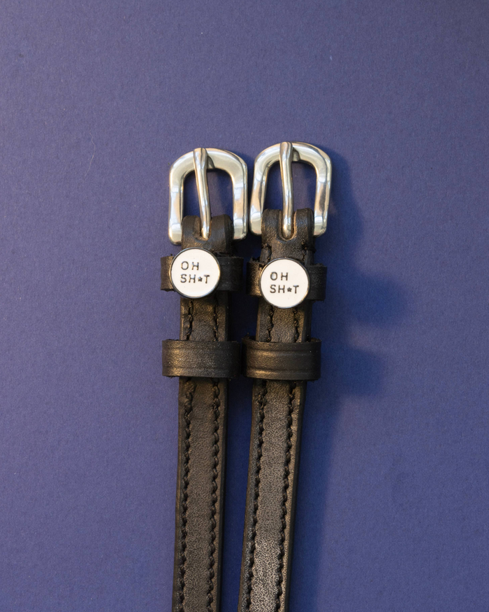 Black leather OH SH*T charm spur straps flat-laid on a navy background