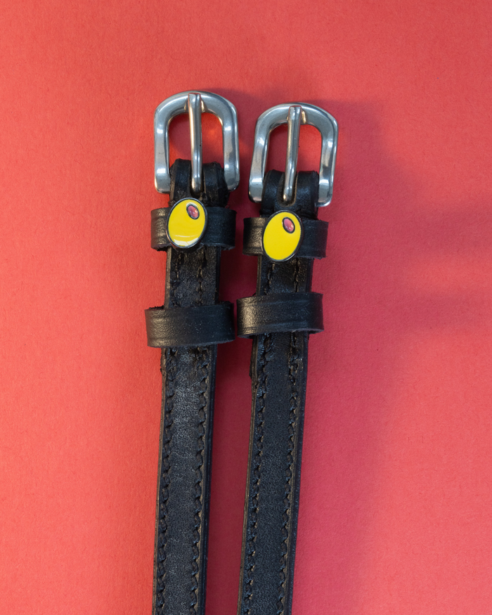 Black leather ManeJane spur straps with green olive charms on a coral red background