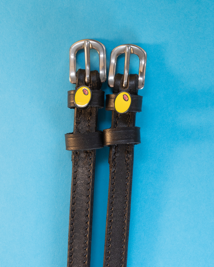 Black leather olive charm spur straps flat-laid on a light blue background