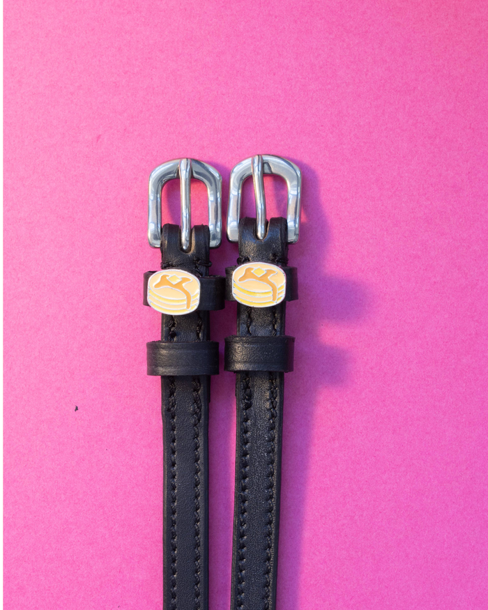 Black leather ManeJane pancake spur straps with silver buckles, flat-laid on a pink background
