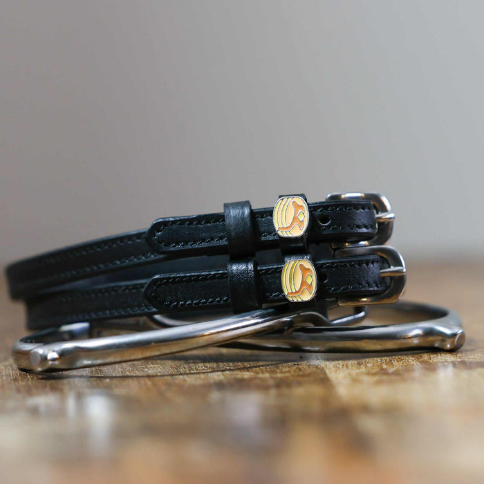 Black leather pancake spur straps coiled beside a stirrup on a wooden surface