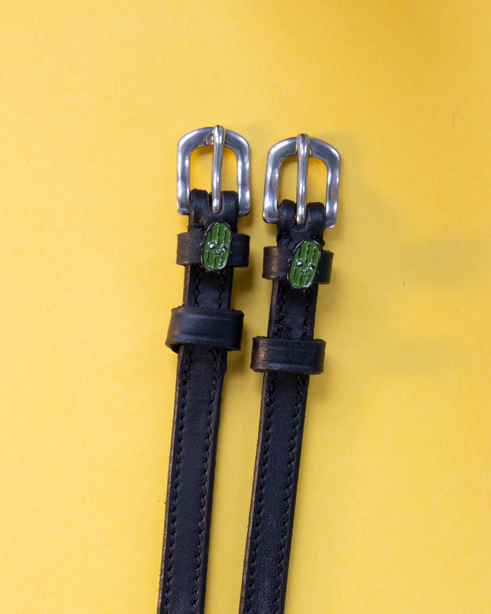 Pair of black leather pickle spur straps with silver buckles laid flat on yellow