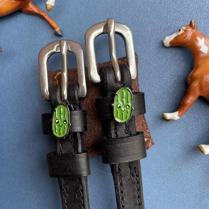 Green pickle charm spur straps with toy horses on blue, ManeJane lifestyle flat-lay