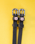 Black leather ManeJane spur straps with pink pig-face charms on a yellow background