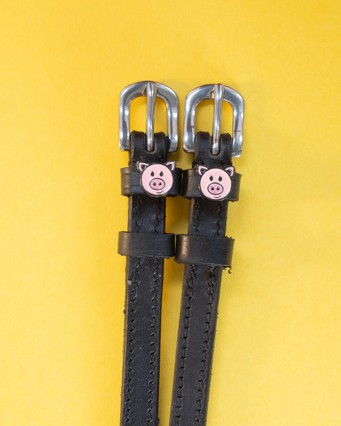 Black leather ManeJane spur straps with pink pig-face charms on a yellow background