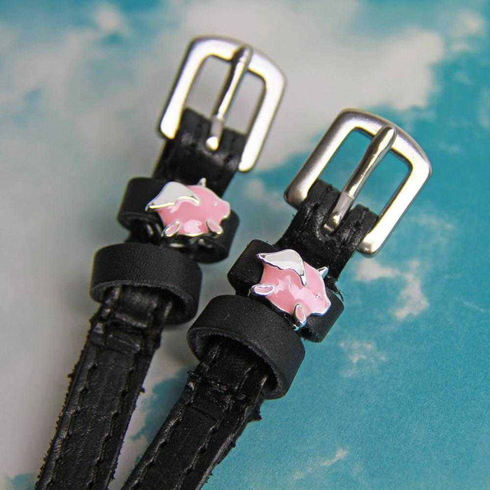 Angled close-up of winged pig charms on ManeJane spur straps against a cloudy sky