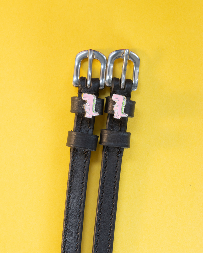 Black leather ManeJane spur straps with silver buckles and pink dinosaur charms on yellow