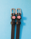 Black leather ManeJane spur straps with pink rhinestone flower charms on a blue background