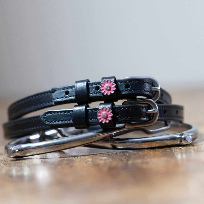 Pink flower charm spur straps coiled beside a stirrup on a wooden tabletop