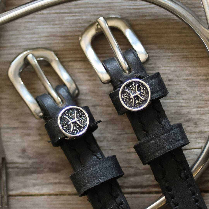 Close-up of Pisces zodiac charms and silver buckles on black spur straps, wood