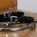 Plain black leather ManeJane spur straps with no charm, coiled by a stirrup on wood