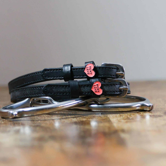 Pony Love heart-charm spur straps coiled beside a stirrup on a wooden tabletop
