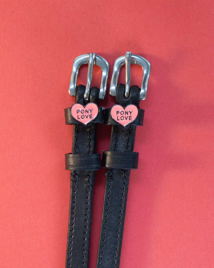 Pair of Pony Love spur straps with pink heart charms laid flat on a coral background