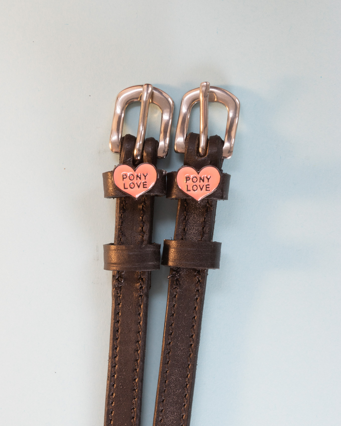 Brown leather ManeJane Pony Love spur straps on a soft blue-grey background