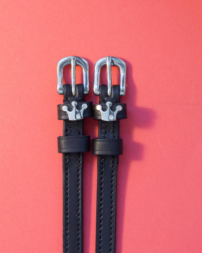 Black leather ManeJane spur straps with silver crown charms on a coral background